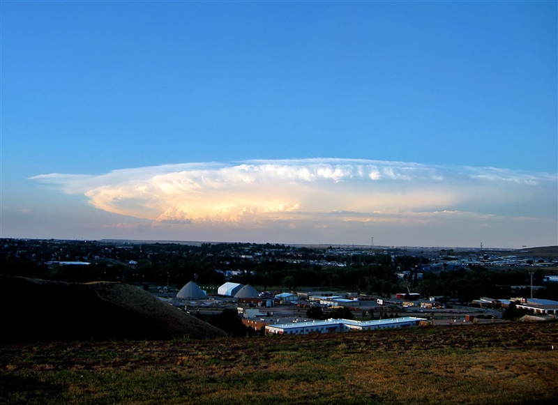 This image depicts a supercell above buildings. Its sprawling, white clouds are thick with moisture.