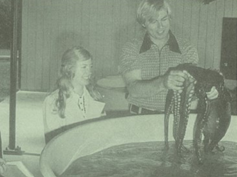 archival black and white photo of a researcher holding an octopus above a large circular water tank while another smiling researcher observes