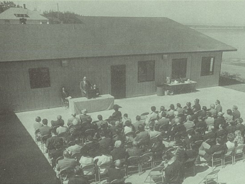 archival black and white photo showing a person speaking from a podium to a seated crowd of spectators in an outdoor courtyard with water in the background