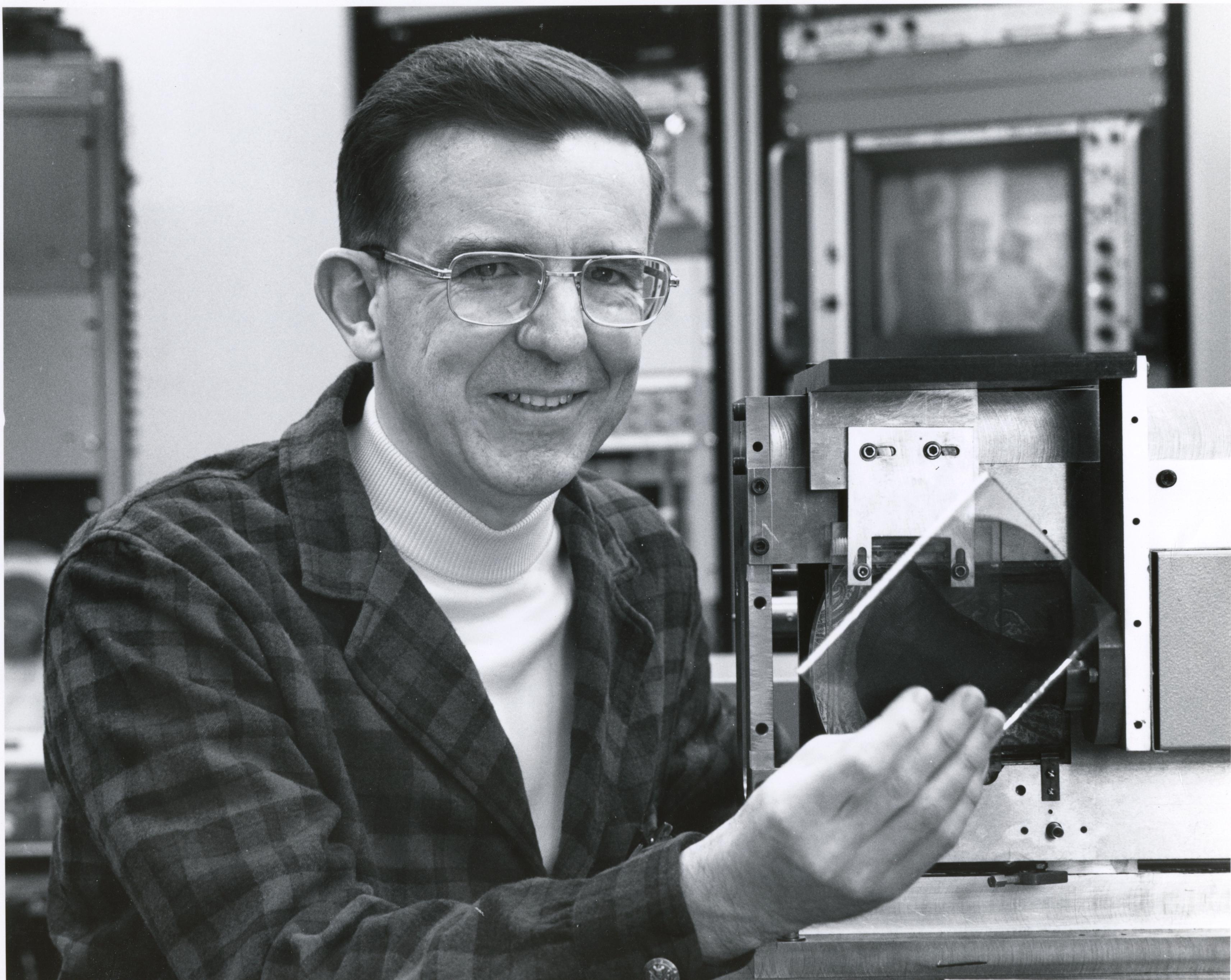 archival black and white photo of Jim Russell, wearing a turtleneck shirt, plaid button-down and glasses, holding the material that would go on to be used for recording music on compact discs
