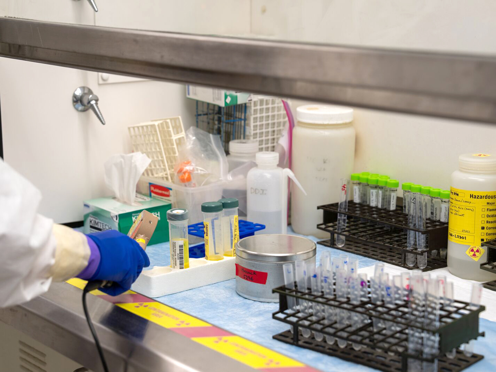A small table area containing samples of pretreated, radioactive tank waste. An arm covered in safety lab-wear and a glove holds a tool, reaching for a container with it.