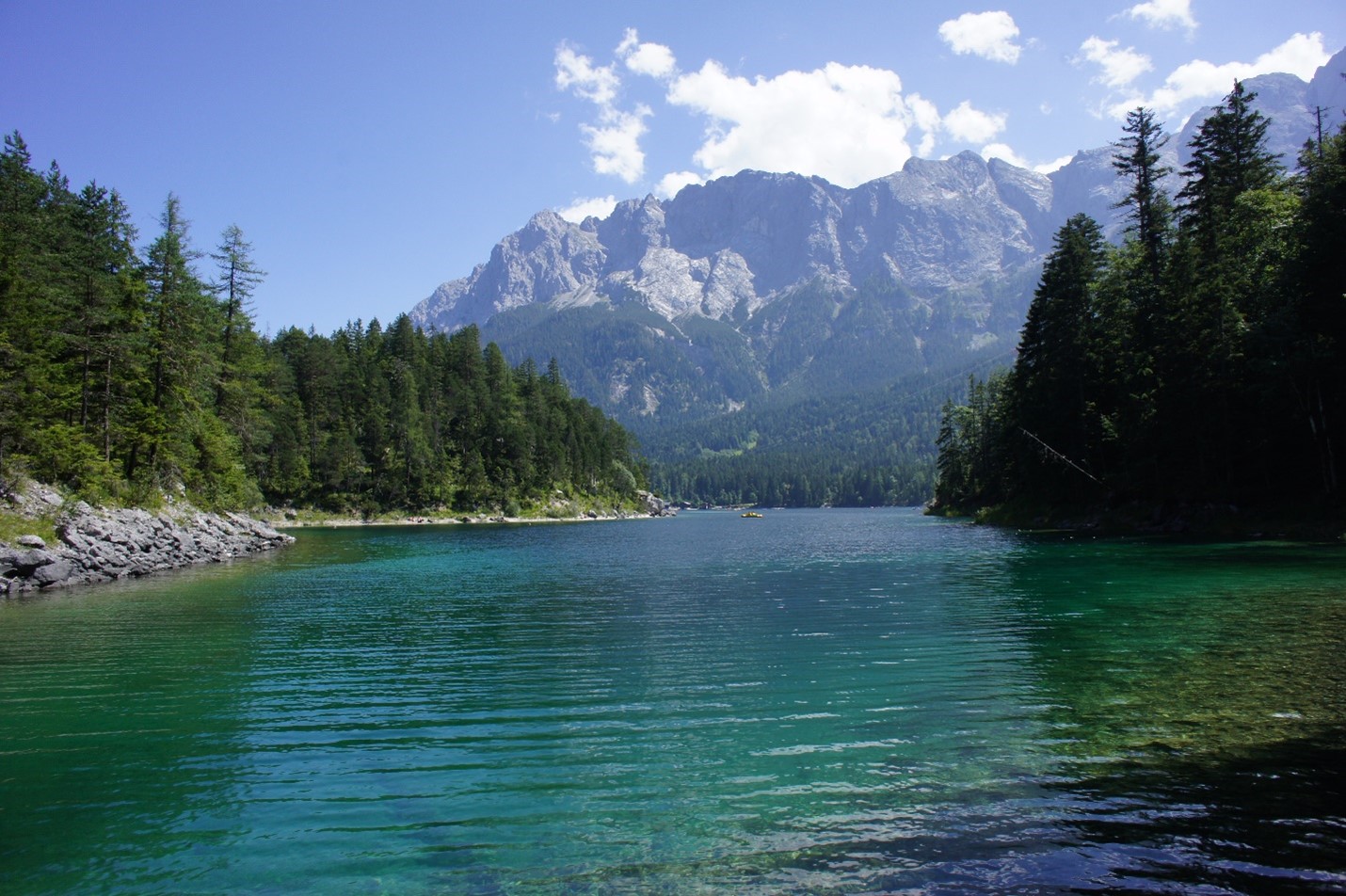 A photograph of a beautiful jewel colored lake surrounded by trees, with a background of mountains