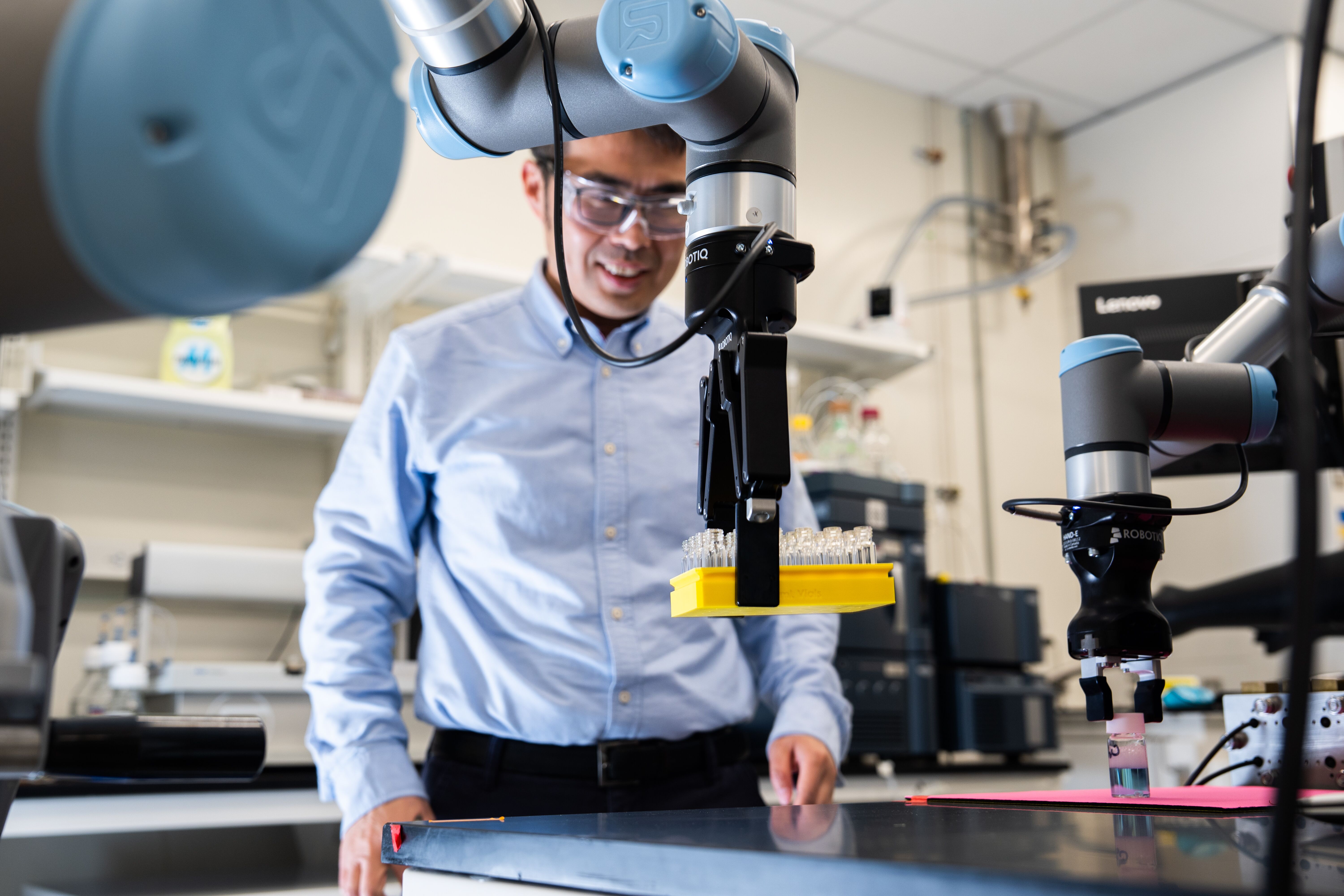 A researcher wearing safety glasses operates a robotic arm holding a tray of small containers.