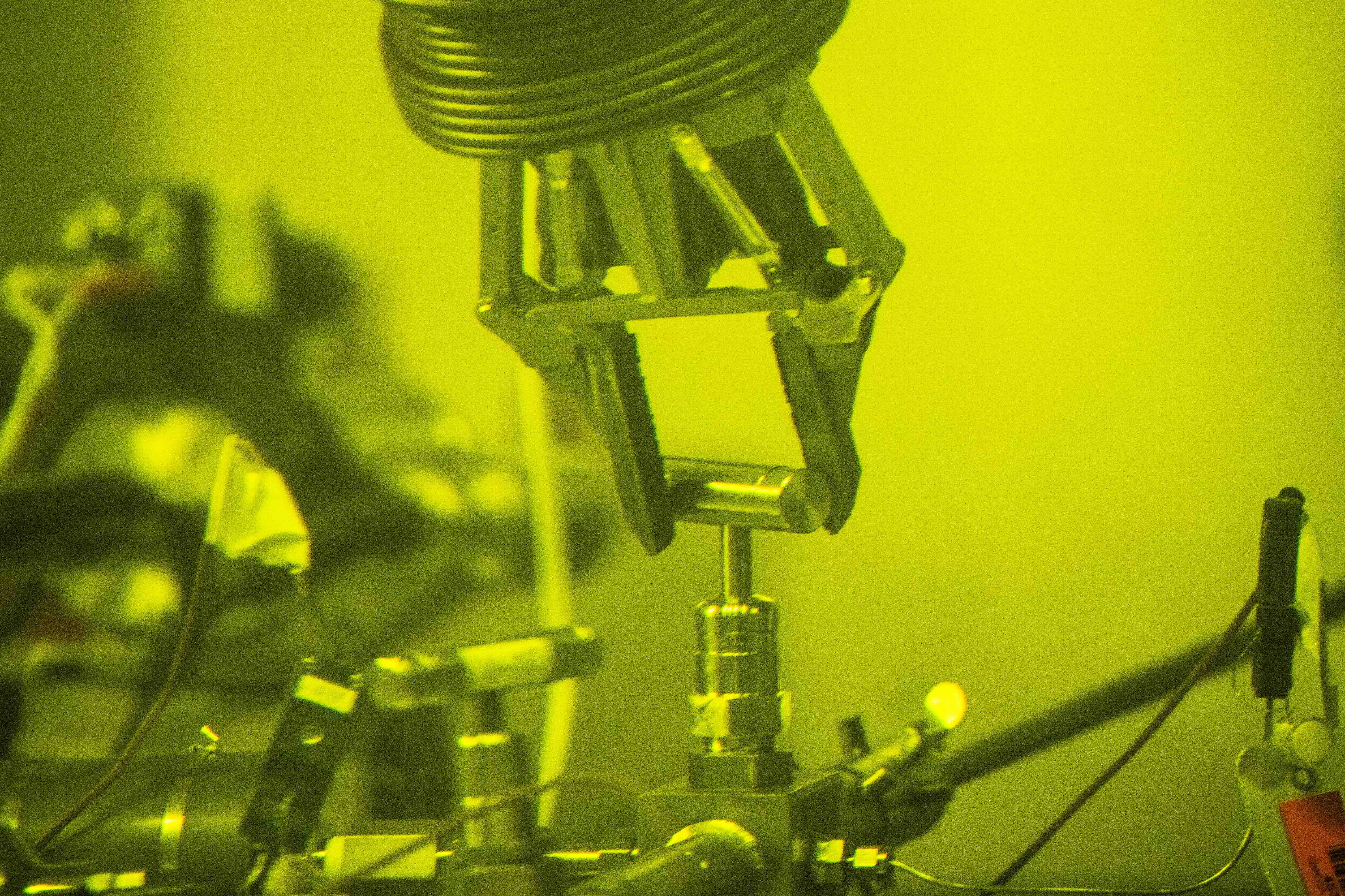 A robotic arm clasps a metal handle, spinning it clockwise so that it may puncture the casing of a nuclear fuel rod inside a hot cell.