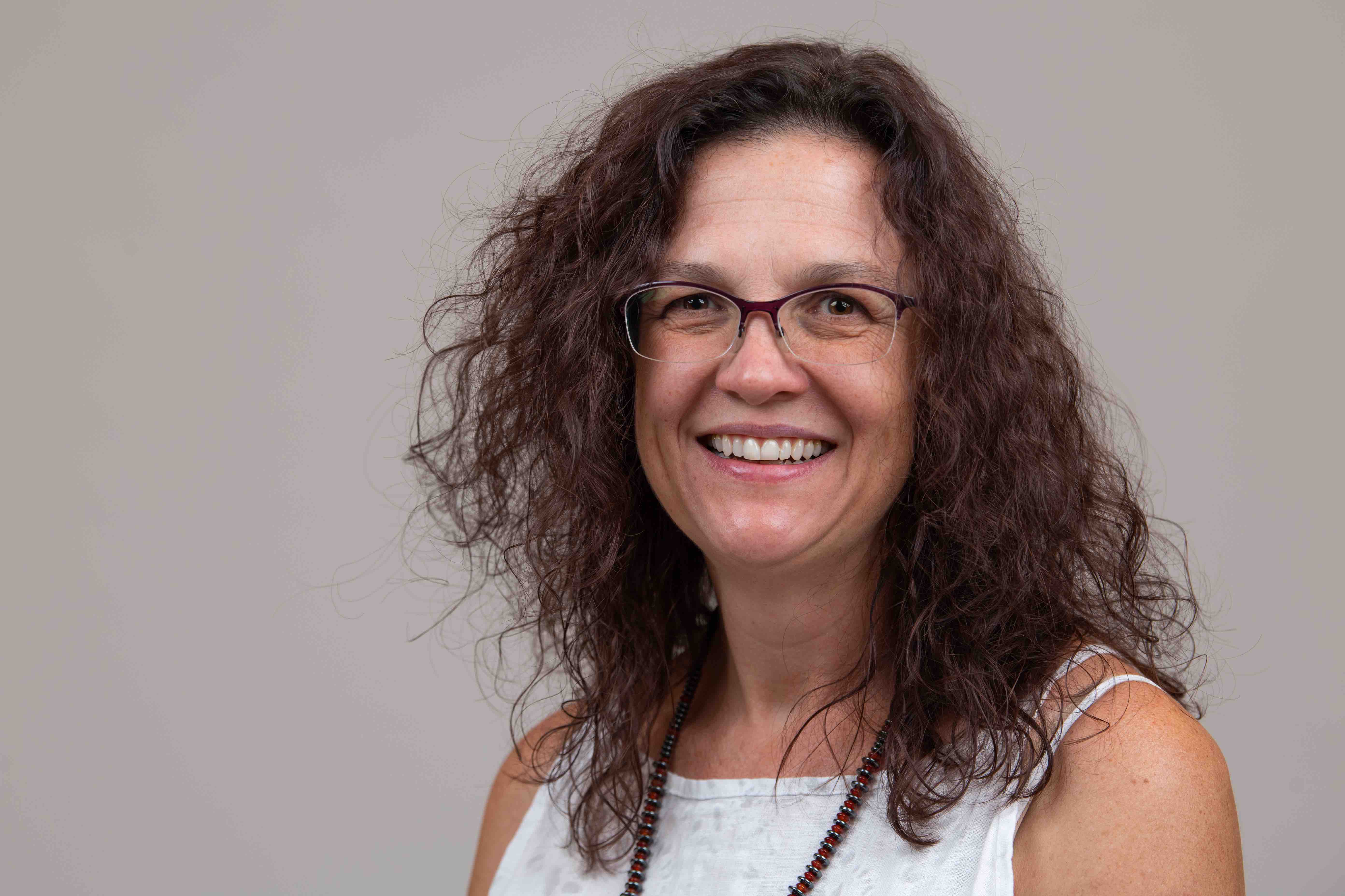 Aurora Clark is a University of Utah professor. She has a big smile, glasses, shoulder-length brown curly hair and is wearing a white sleeveless dress.