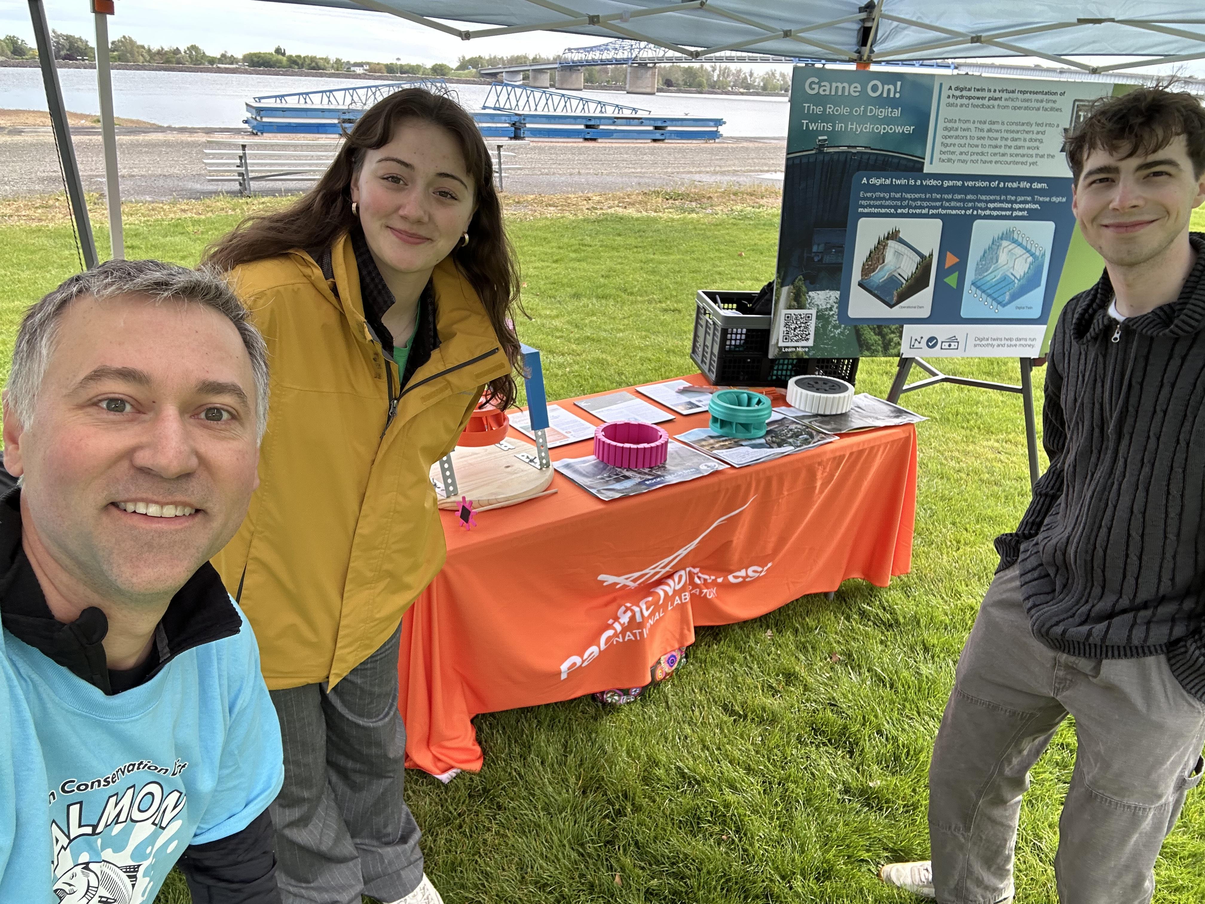 researcher and two college students stand in front of a demonstration table for explaining the digital twins project to students