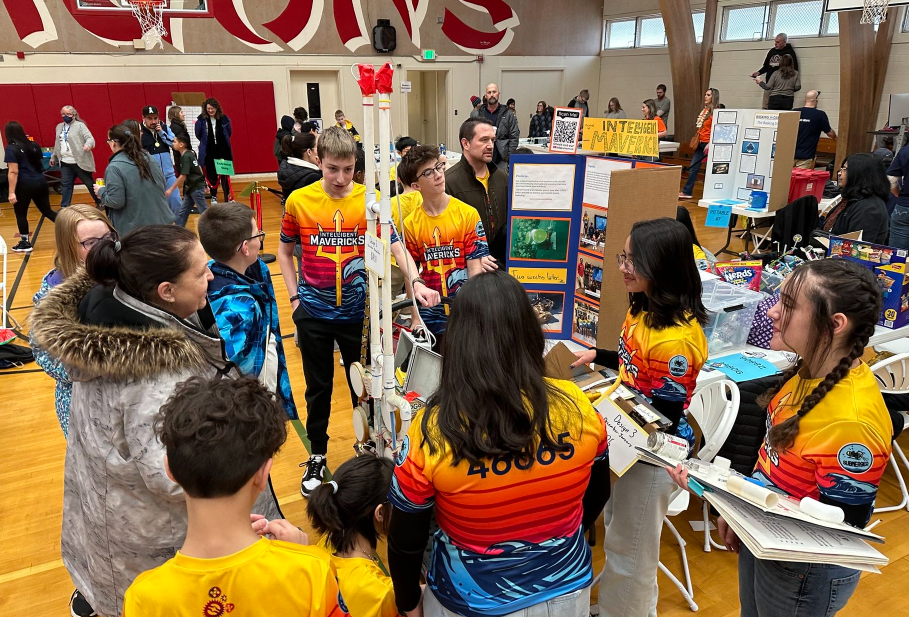 The Intelligent Mavericks team clustered around their table at a competition, explaining their poster board and robot to a competition attendee