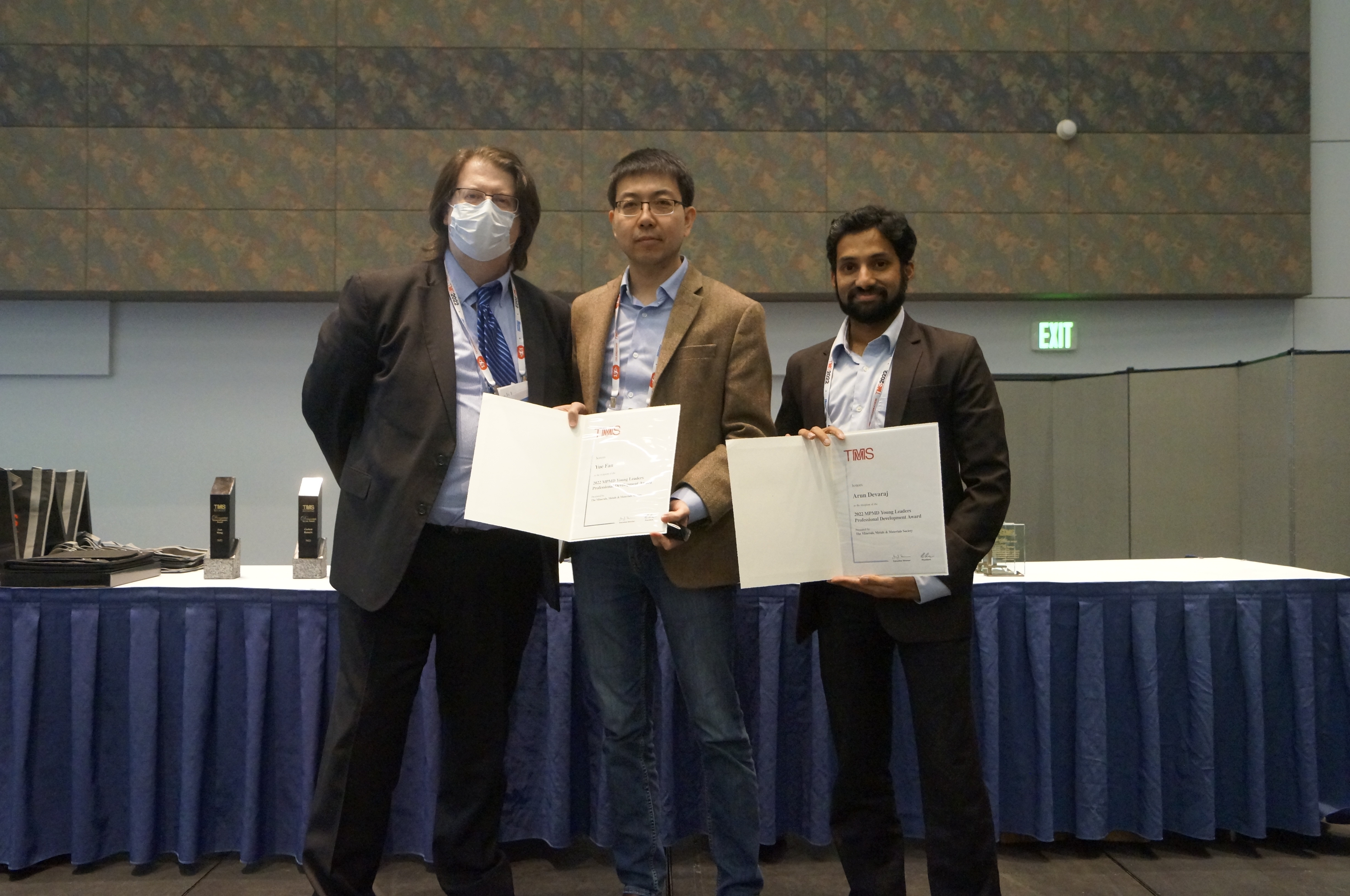 Three men stand side by side, two displaying folders with award certificates