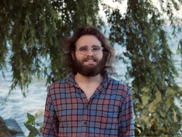 A person standing outdoors with leaves and ocean in the background.