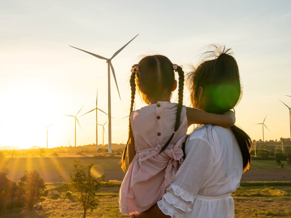 Photo of a person holding a child, looking towards an open field that contains wind turbines.