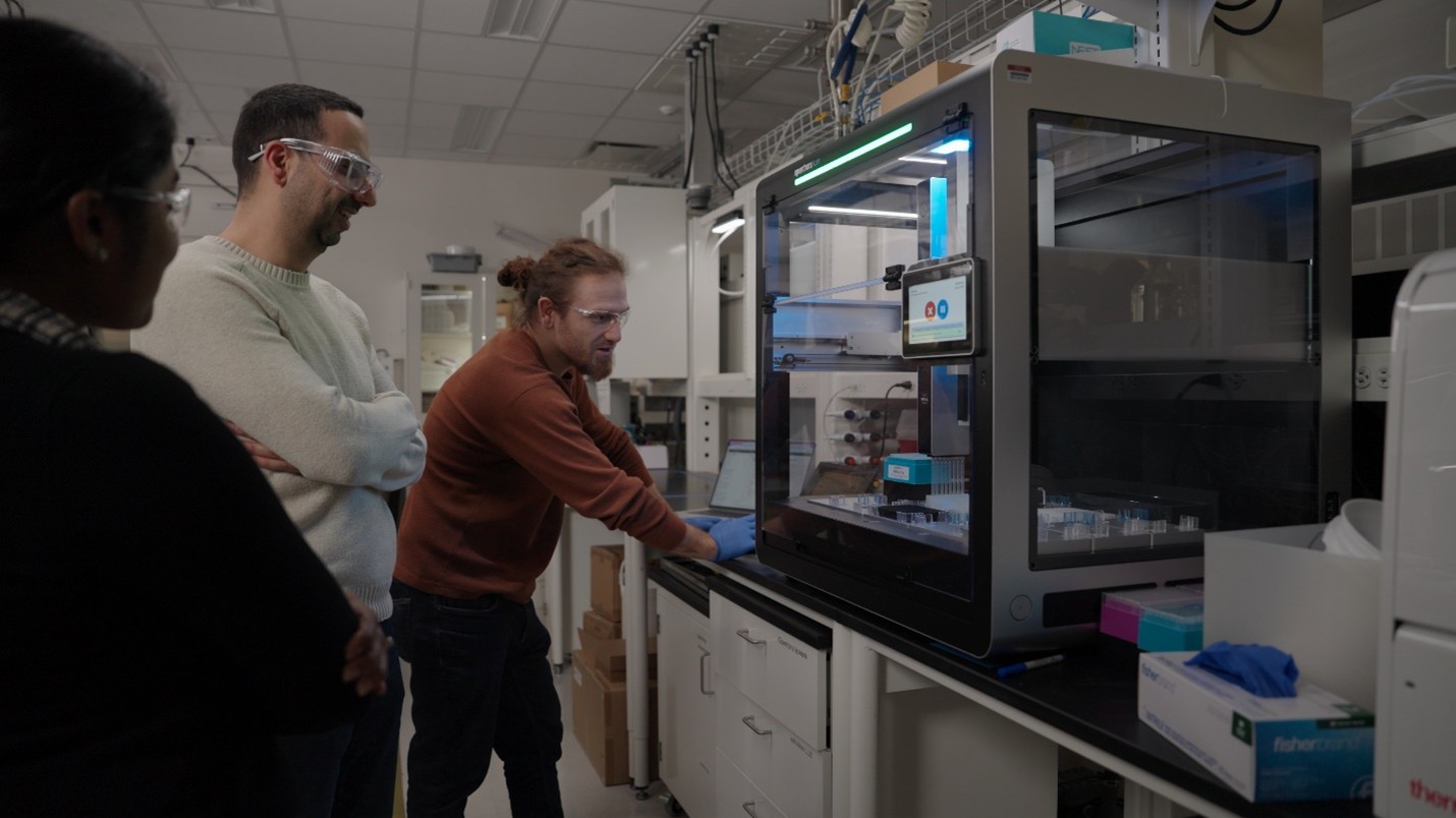 Three researchers wearing safety goggles operating an enclosed machine.