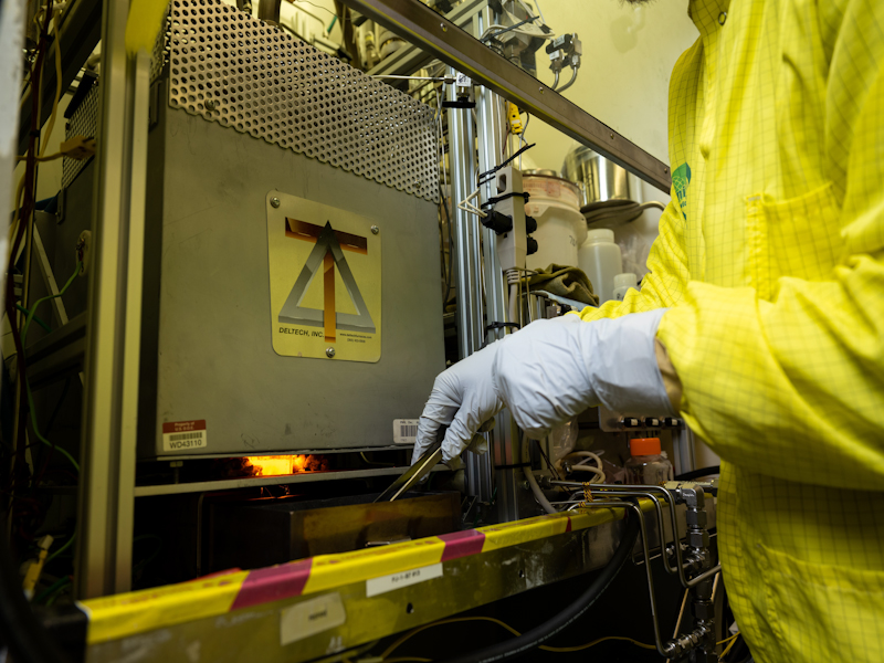 Photo of a person wearing safety clothing and gloves holding a pair of tongs inside the Radiochemical Processing Lab.