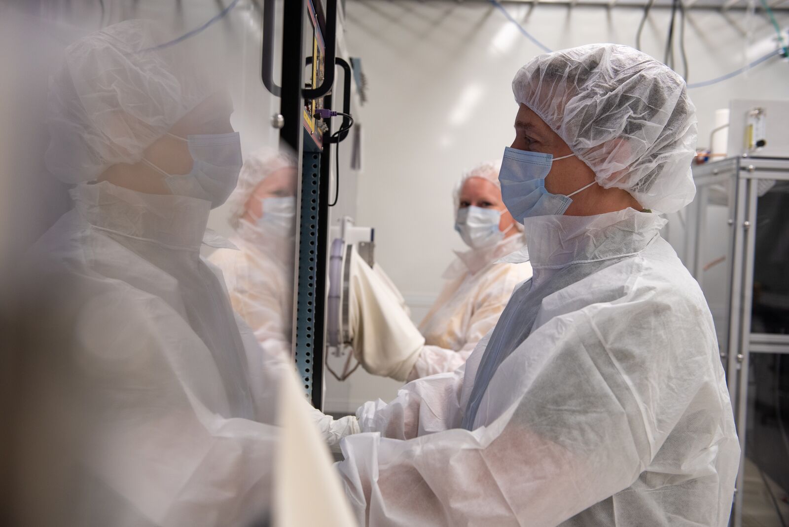Two scientists wearing protective laboratory coats and other gear extend their arms into a laboratory glovebox.