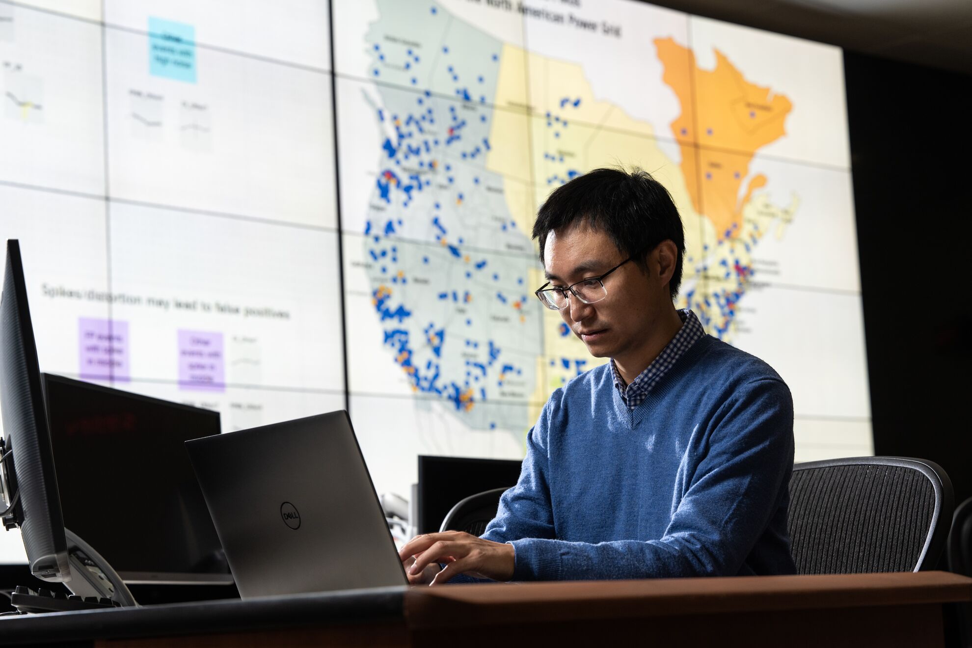 A PNNL researcher using a computer to apply artificial intelligence measurements to the electrical grid; an image of the U.S. electrical grid is in the background.