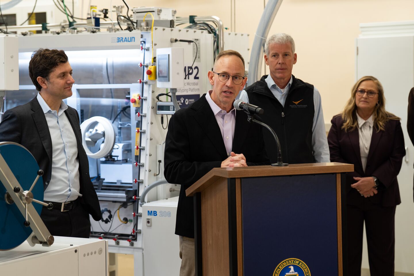 A PNNL researcher speaks at a podium with Secretary of Energy Chris Wright and PNNL Lab Director Deb Gracio in the background.