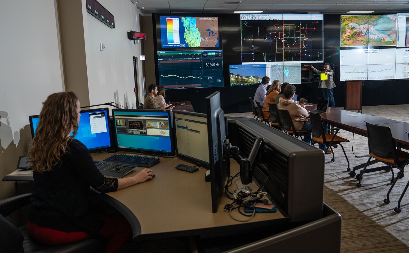 Photograph shows people sitting in a conference room looking at various screens, large and small.