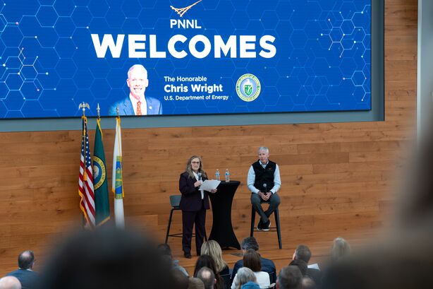 a distant view of two professionals sitting on a stage beneath a digital display screen that reads "welcome"
