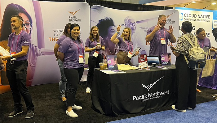Photo of PNNL staff members standing at a 2023 Grace Hopper Celebration recruitment booth, smiling at the camera.