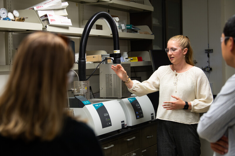 Madeline Bartels stands in front of equipment giving an explanation to visitors in the laboratory.