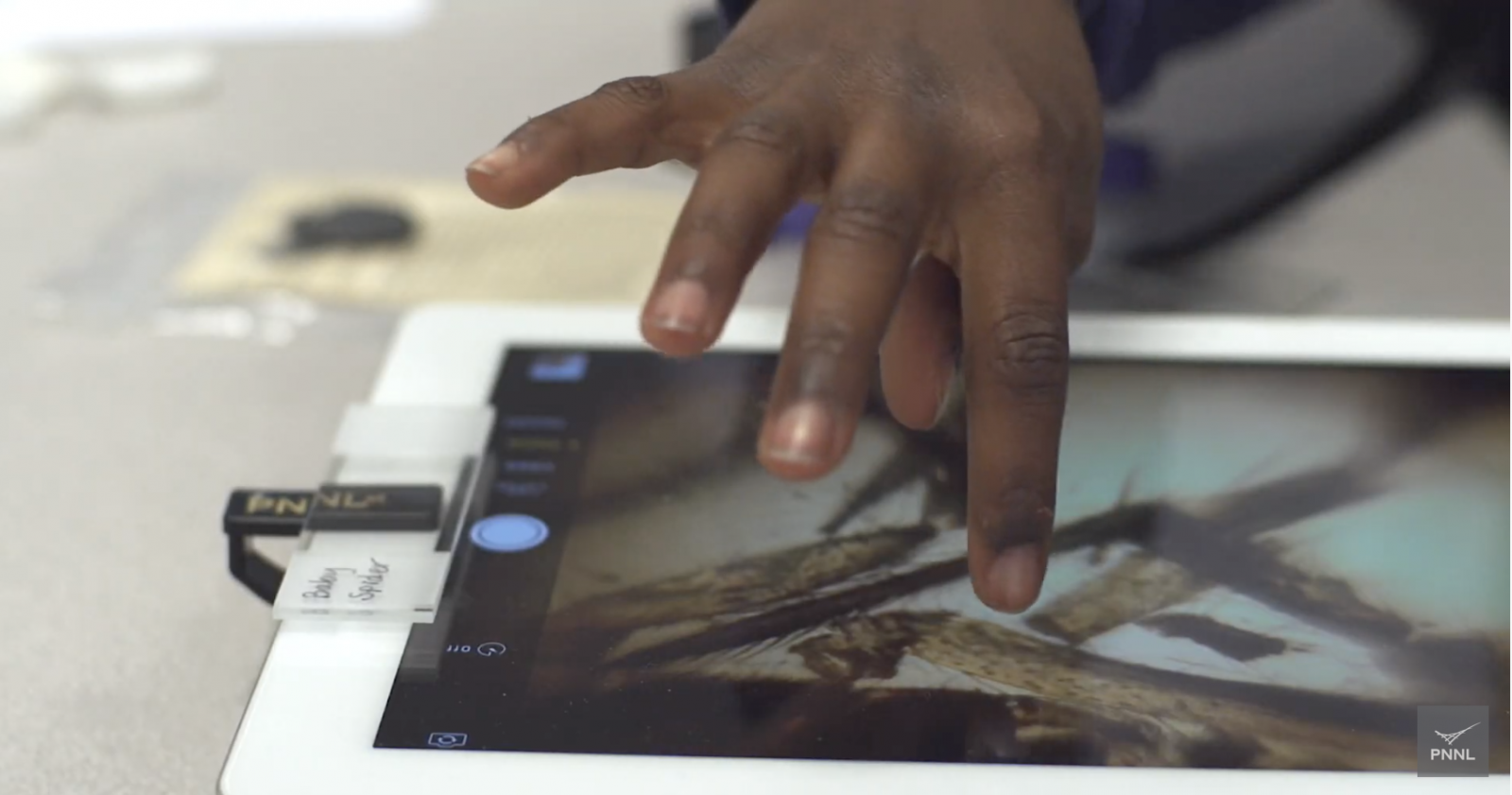 hand touching the screen of a tablet with smartphone microscope clipped to device's camera