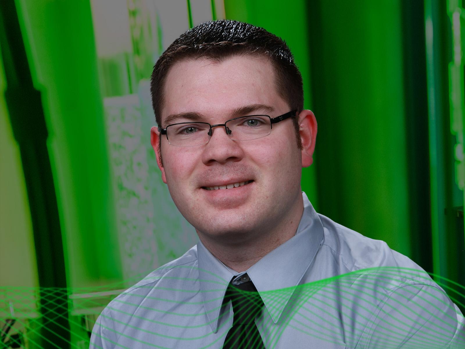 Adam Roberts wearing a collared shirt and tie in front of a green background