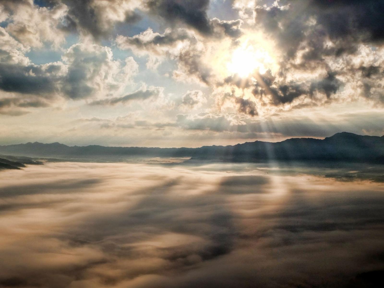 The sun breaking through a clouded sky over a mist-covered Bodega Bay with mountains in the distant background