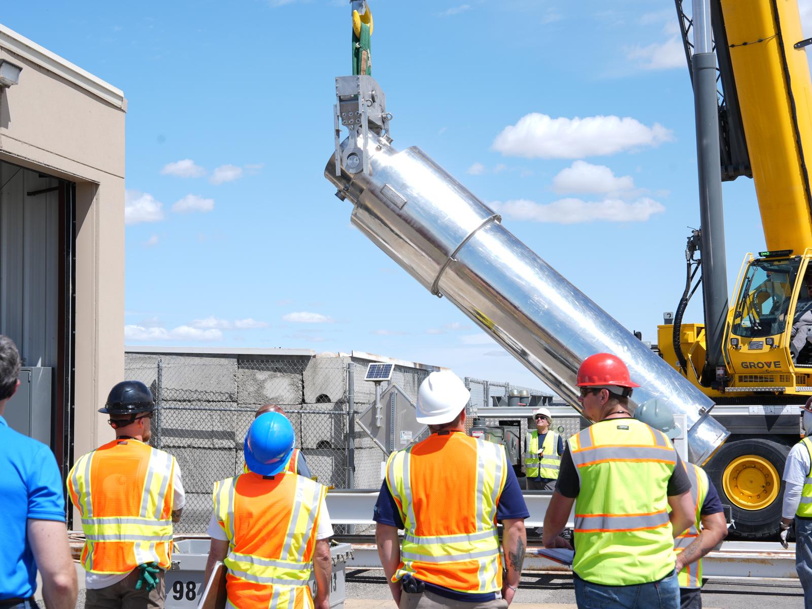 Several people stand before a massive, 30-ton canister containing eleven nuclear fuel rods. The canister is being lowered onto a wheeled track by a crane. 