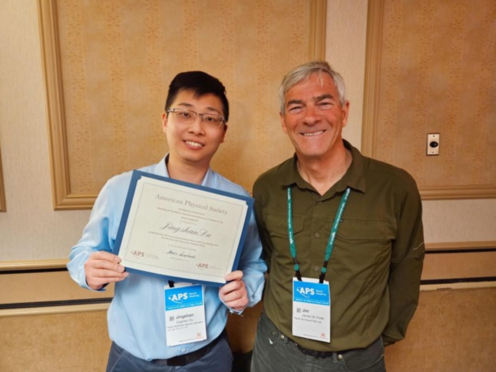 Photograph of two people smiling at a conference. The person on the left is holding an award certificate.