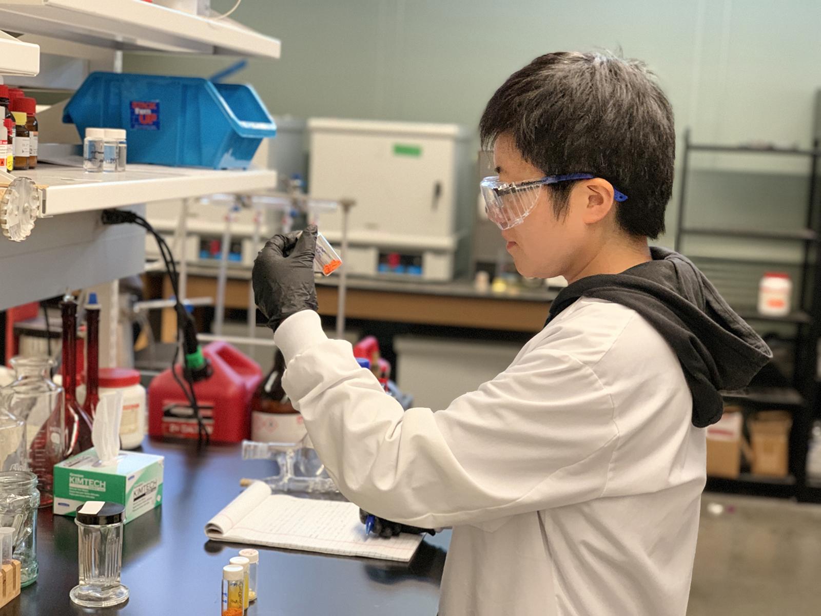 Young scientist in lab coat looks at small jar of materials in a science lab.