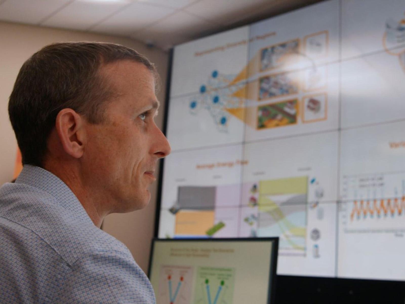 a researcher in a collared shirt in the foreground looking to the right at a large digital screen full of charts and graphs