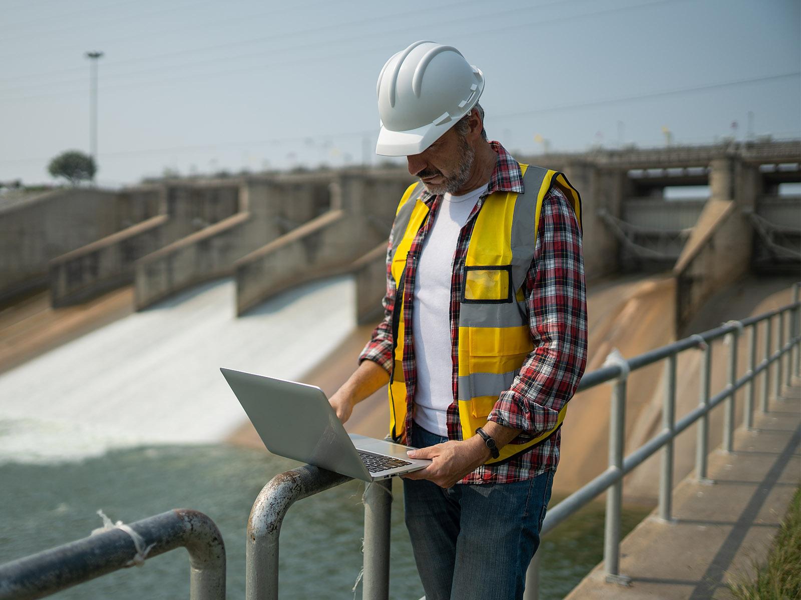 A man with a hardhat looks at a laptop in front of a hydropower dam.