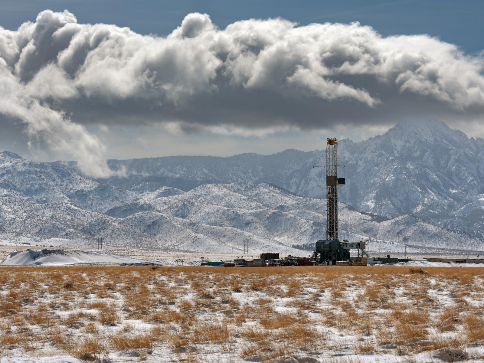 Photograph shows a desert landscape against a mountain range, with large clouds in the sky. In the foreground is a geothermal power plant featuring a tall tower.