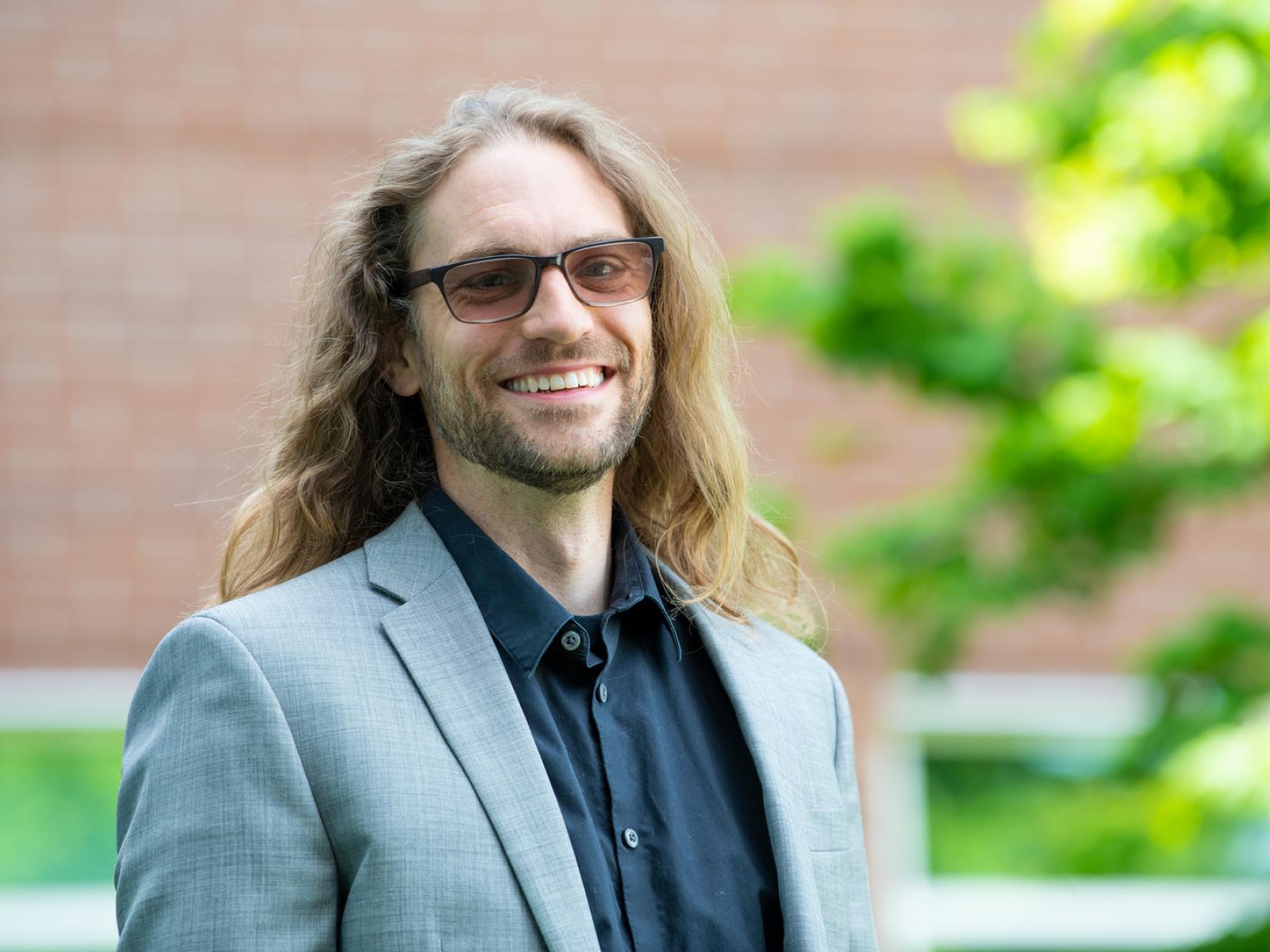 Photograph of a smiling man wearing business attire standing outside in the sunlight