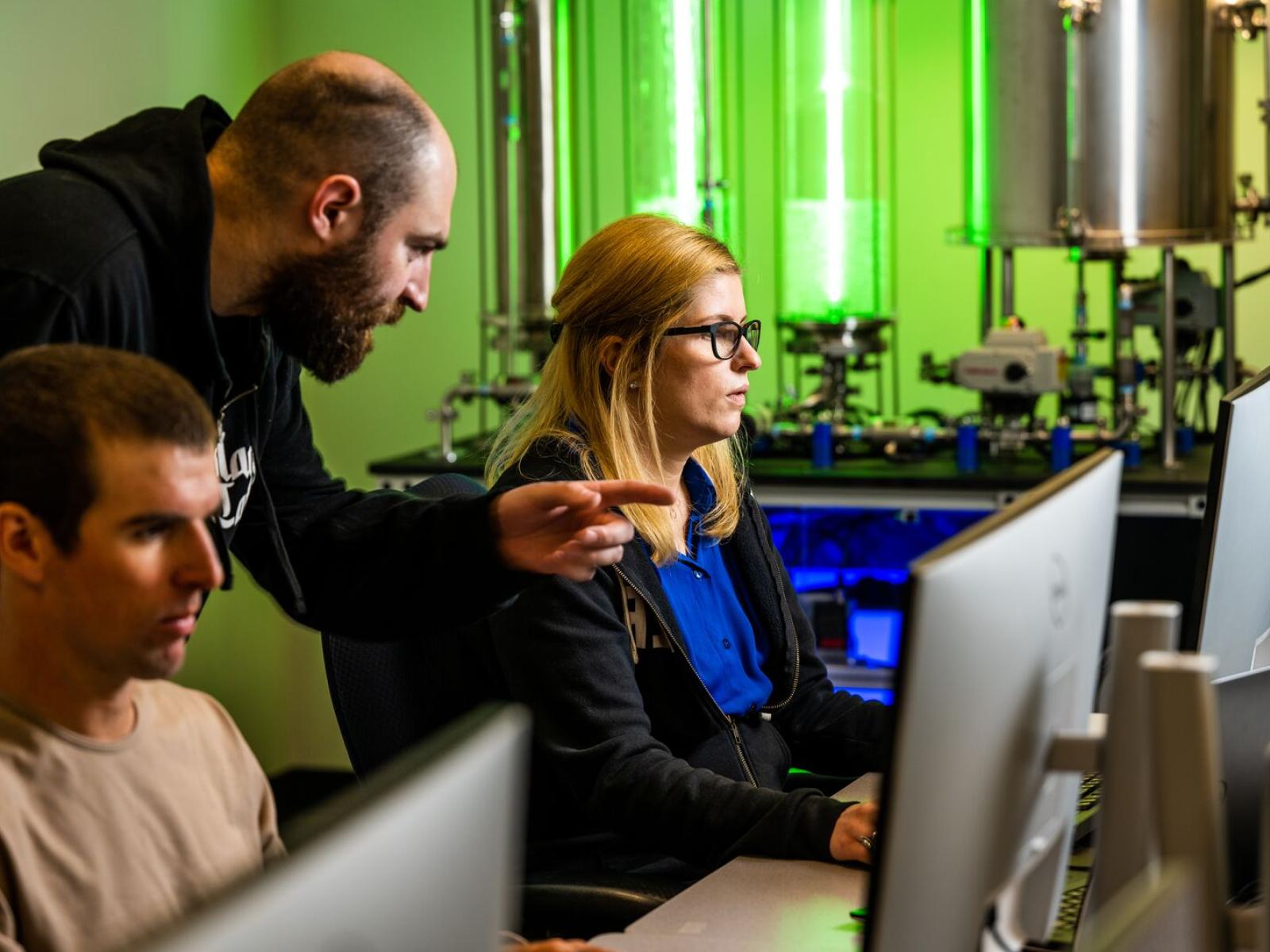 Two people looking at computer screens. In back of them, a third person leans over their shoulder to view a computer screen. In the background there's an instrument mimicking a water treatment plant with two tall glass cylinders filled with water.