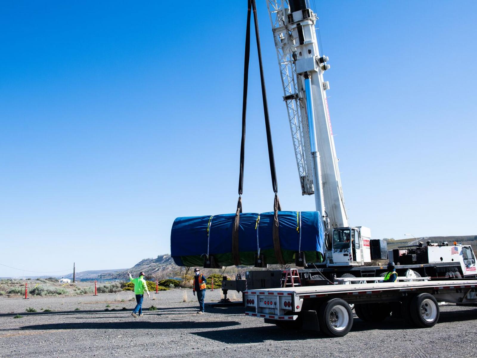 A crane lifts a spent fuel canister, wrapped in a blue tarp, from a flatbed truck.