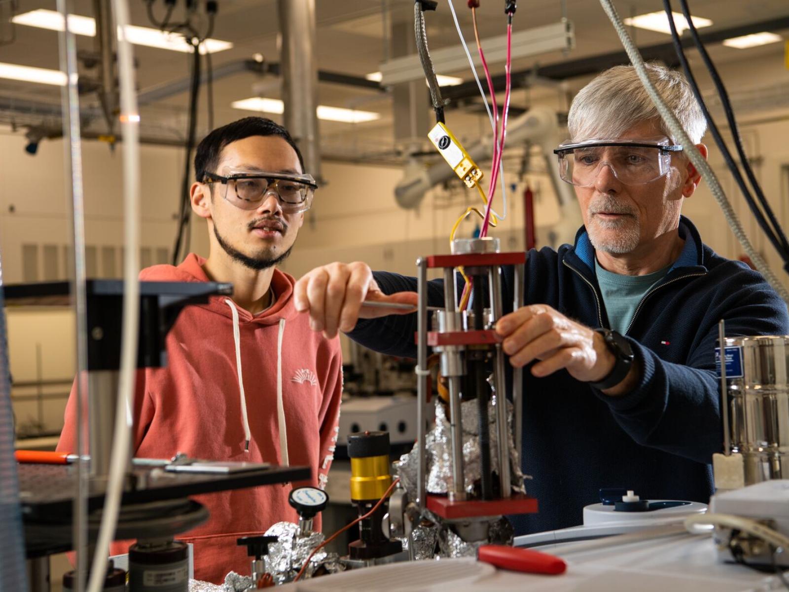 Photo of two researchers in-doors, wearing safety goggles. One works with an apparatus while the other watches.