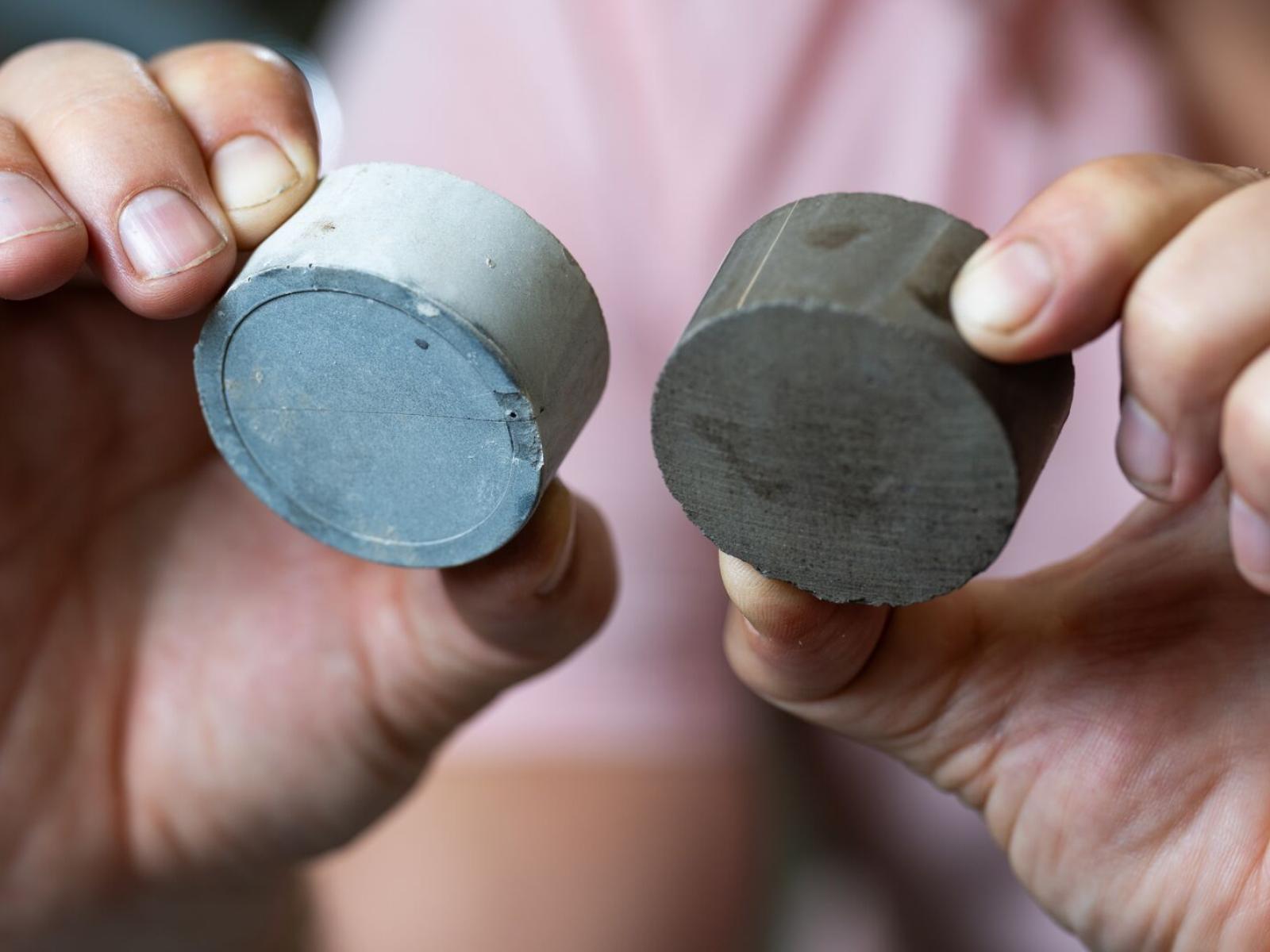 A researcher holds up two samples: on the left, a cylinder of cement made with traditional supplementary materials; on the right, a cylinder of cement made with algal biochar.
