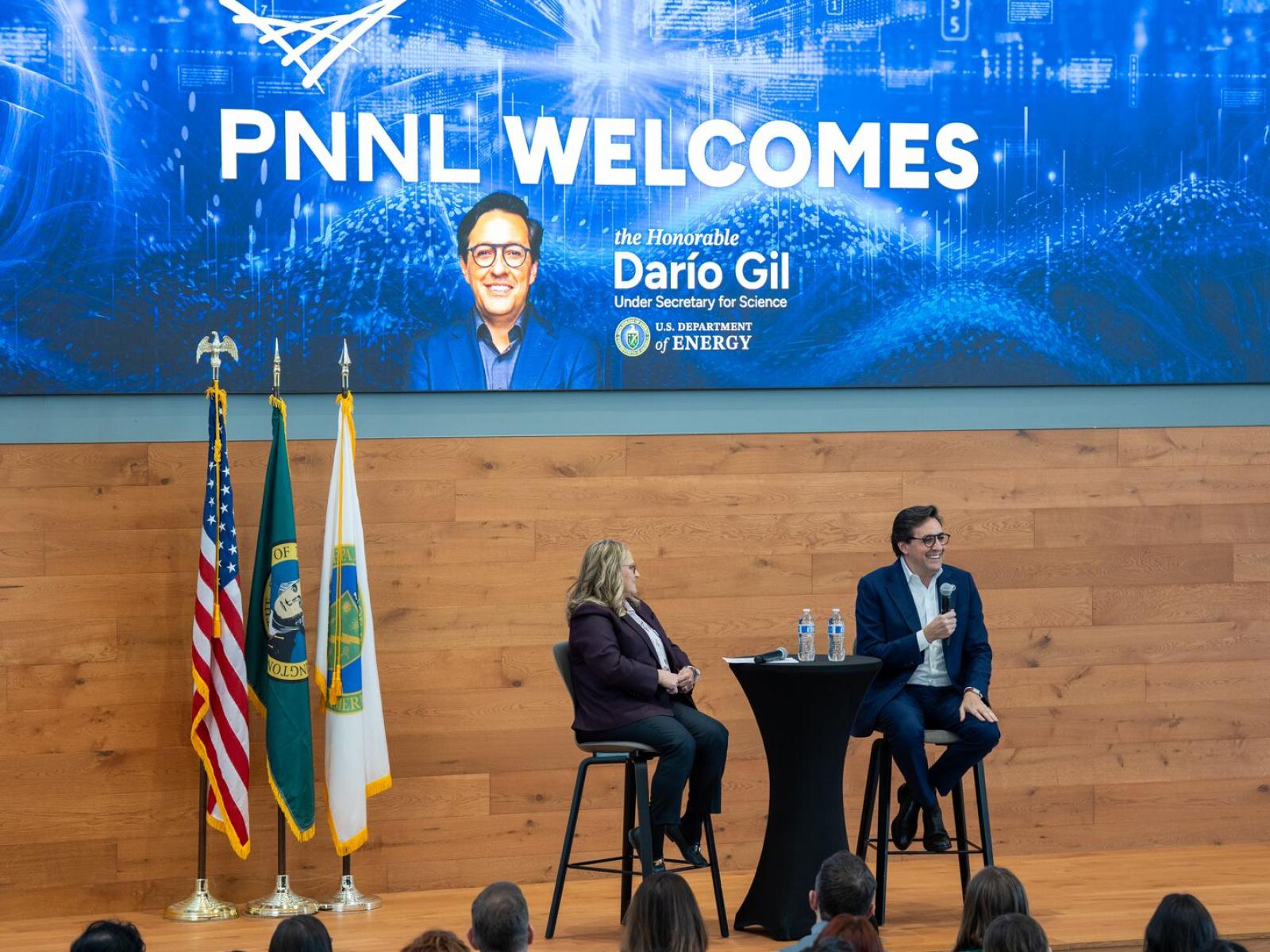 Photo shows a man and a woman sitting on chairs on a stage, holding microphones and looking at an audience. In the background, a projected slide shows "PNNL Welcomes Dario Gil"