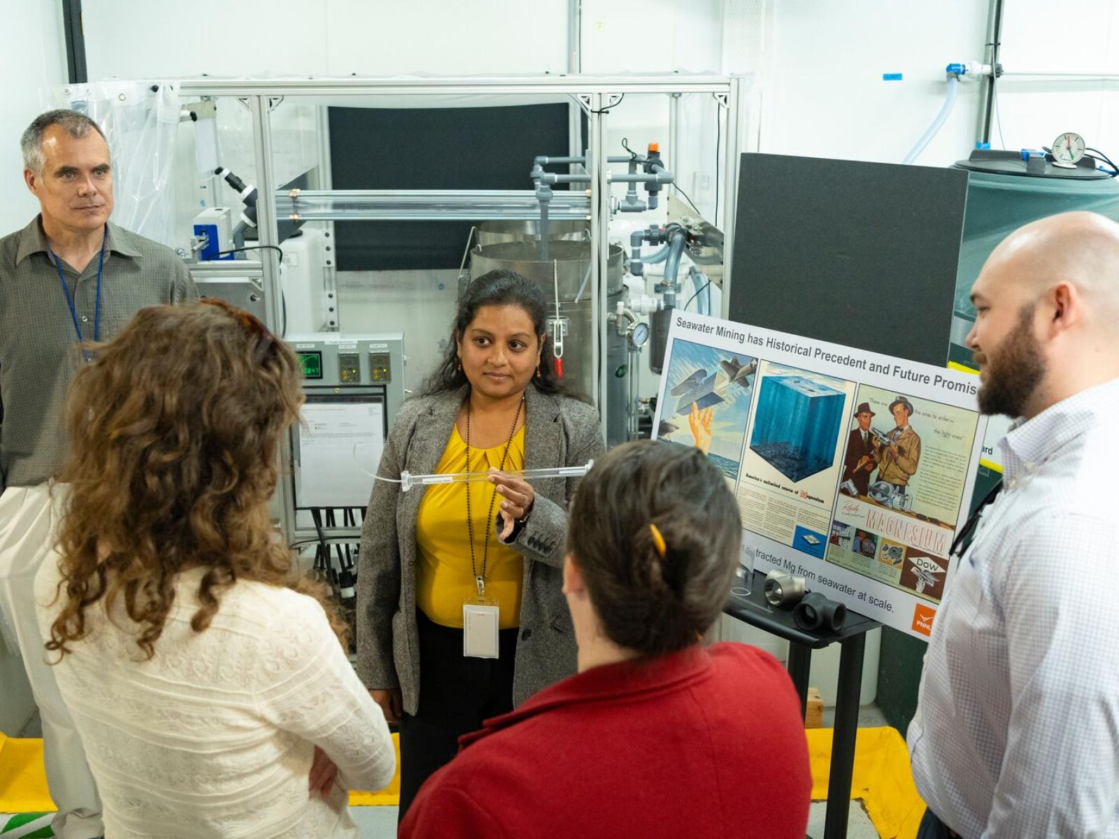 A woman in a grey blazer and yellow shirt holds up a plastic tube from a scientific instrument and shows it to a group of people.