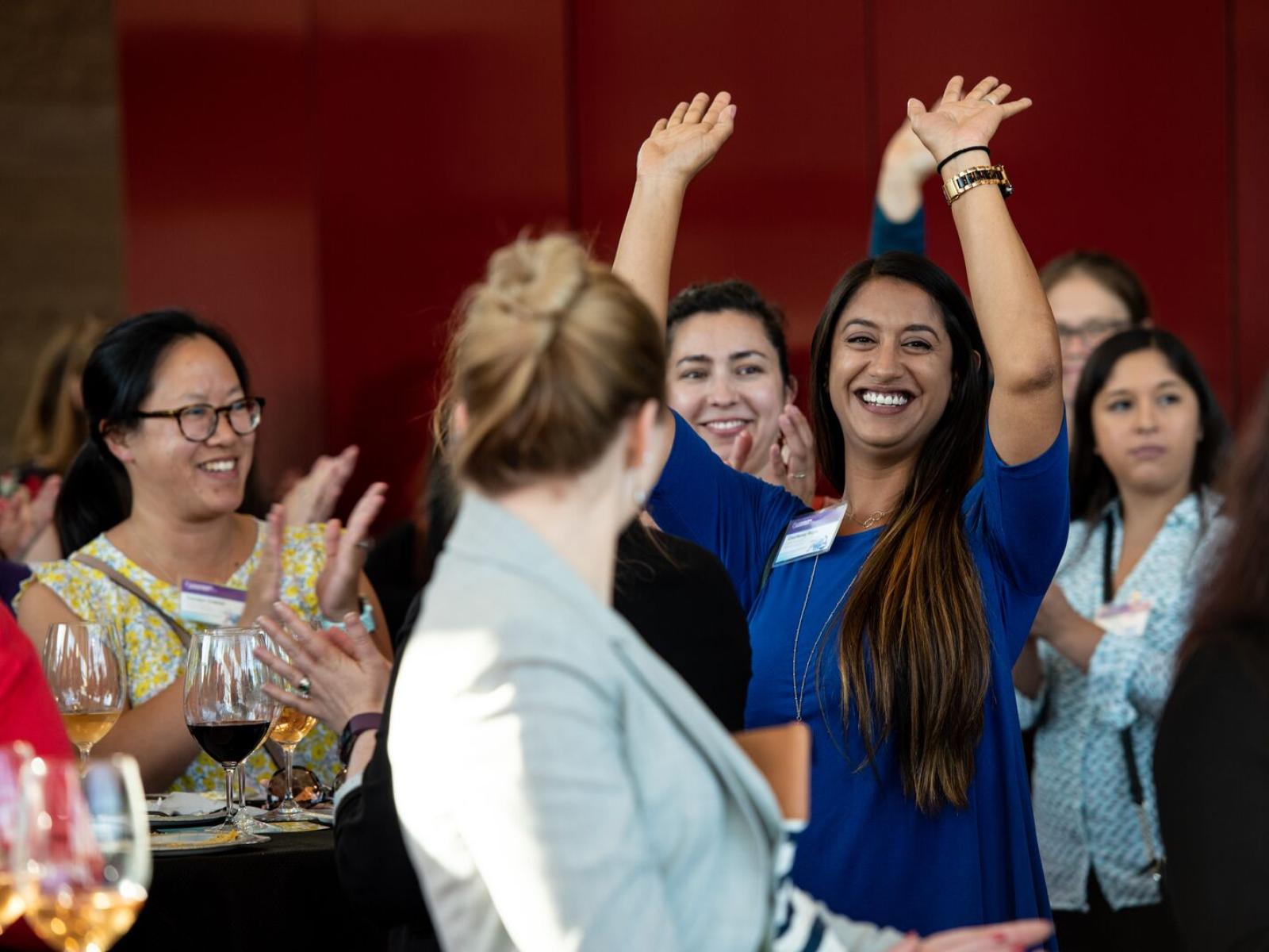 Several women at an event. One women is smiling and waving at the camera.