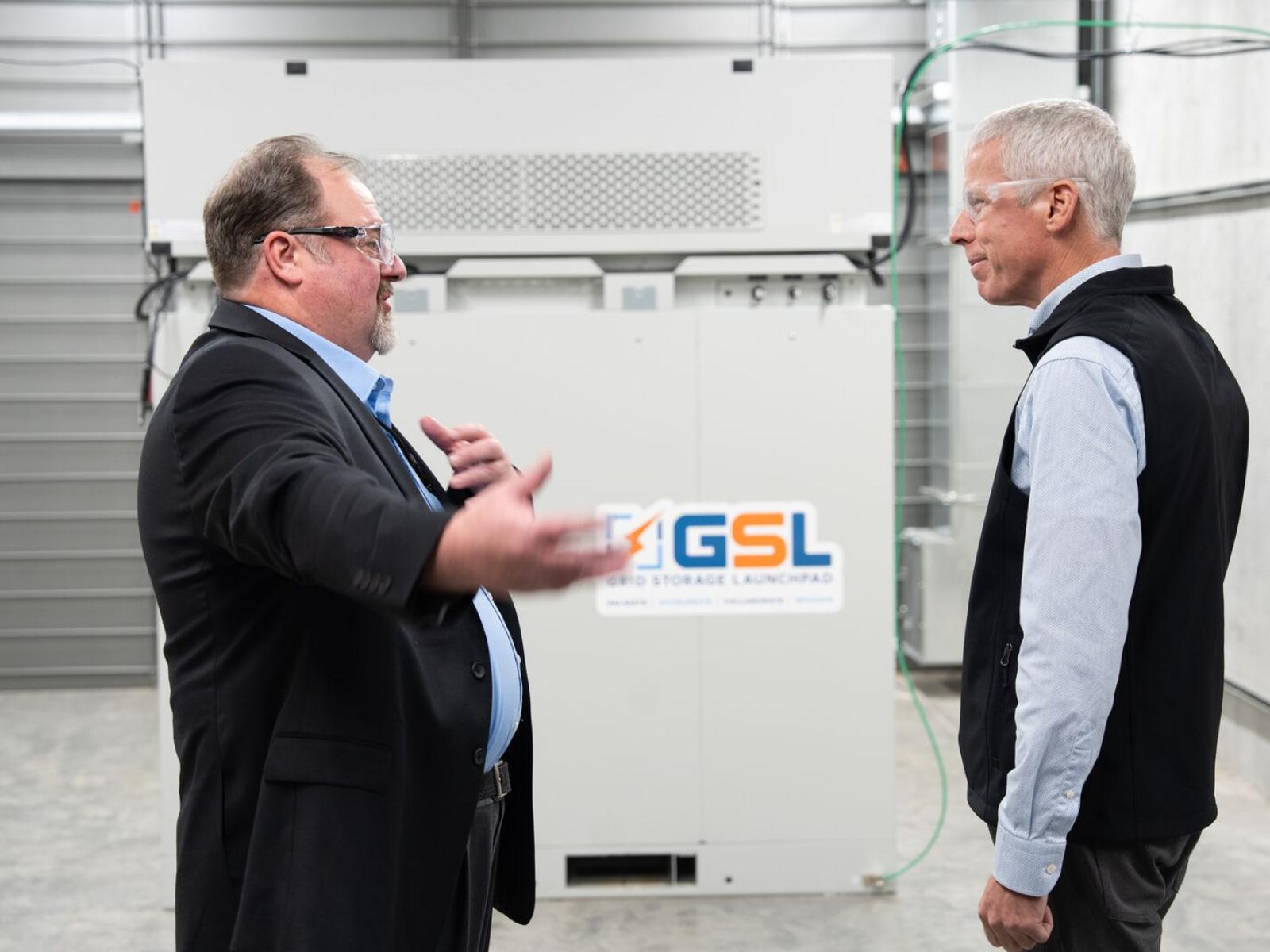 Two men face each other in conversation in front of a large, white battery module, decorated with the Grid Storage Launchpad logo. 
