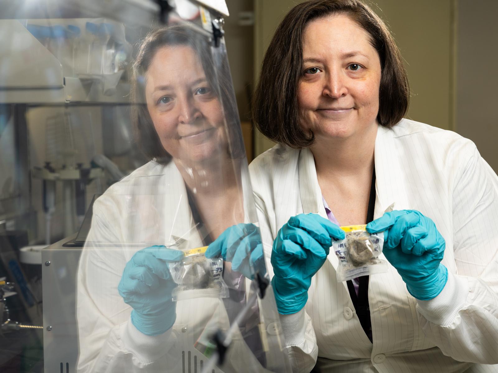 Becky Hess in a lab coat, wearing safety gloves, holding a sample in a small bag.