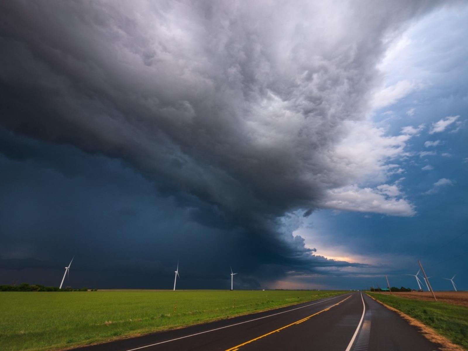 Wall of dark stormclouds above wind turbines, green grass, and a two lane road.