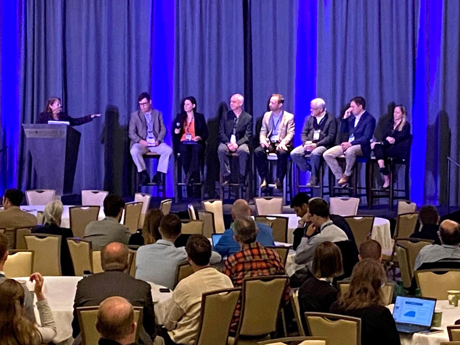 Seven speakers and a moderator on a stage at a conference.