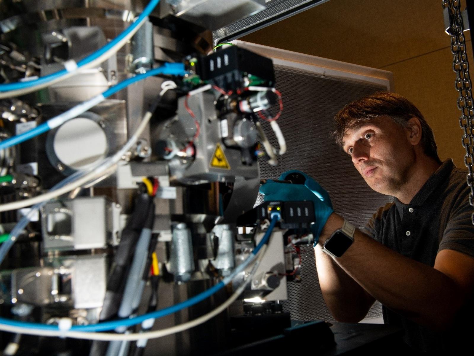 A white man with short brown hair stares intently at an electron microscope as he inserts a sample
