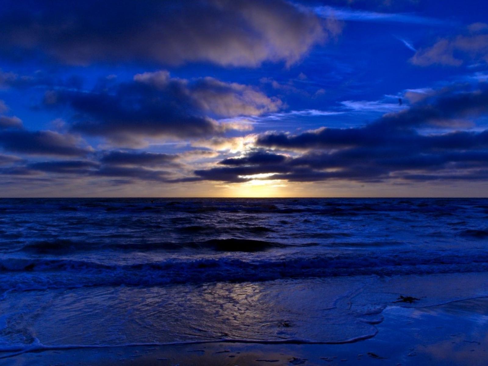 A deep blue hued sunset image above a beach, with the sun setting behind clouds.