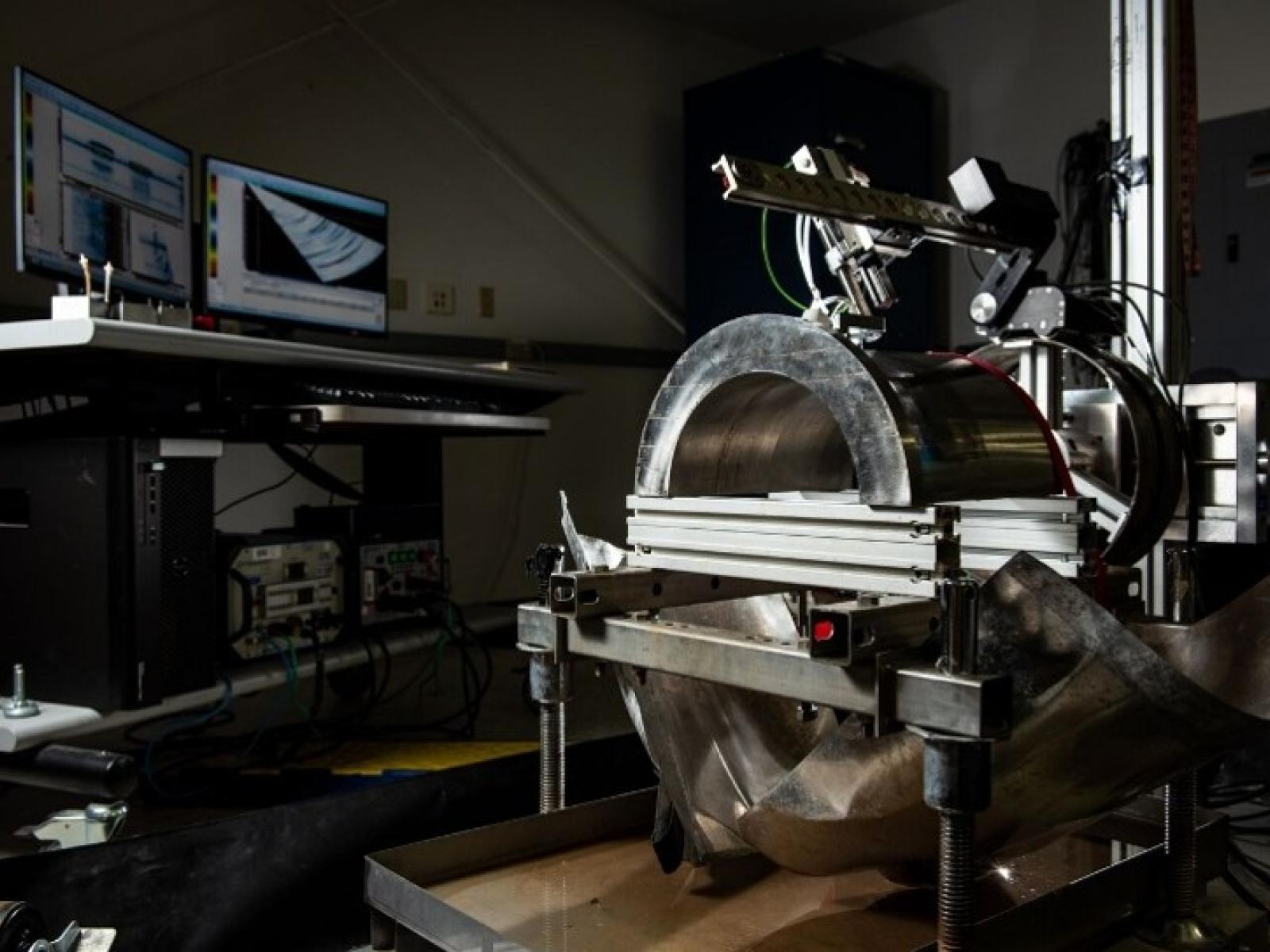 A room with computer screens in the background hooked up to a device that is scanning a large piece of metal material