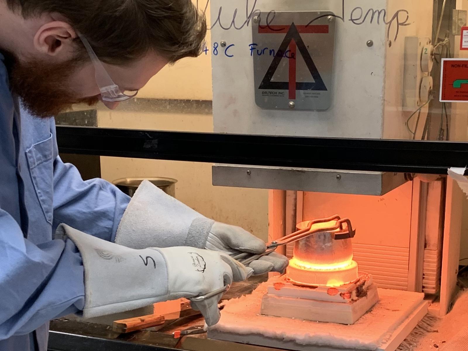 Male researcher wearing thermal gloves uses forceps to grab a crucible of melted glass