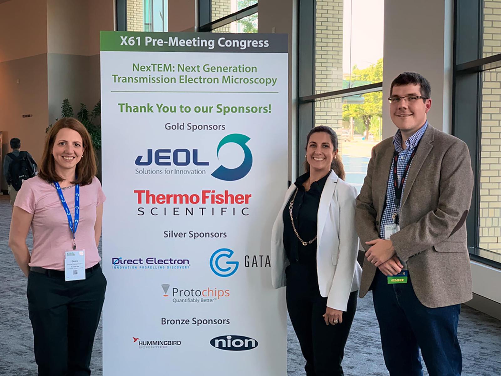 Two female scientists and one male scientist stand by a banner listing the sponsors for the NexTEM workshop.
