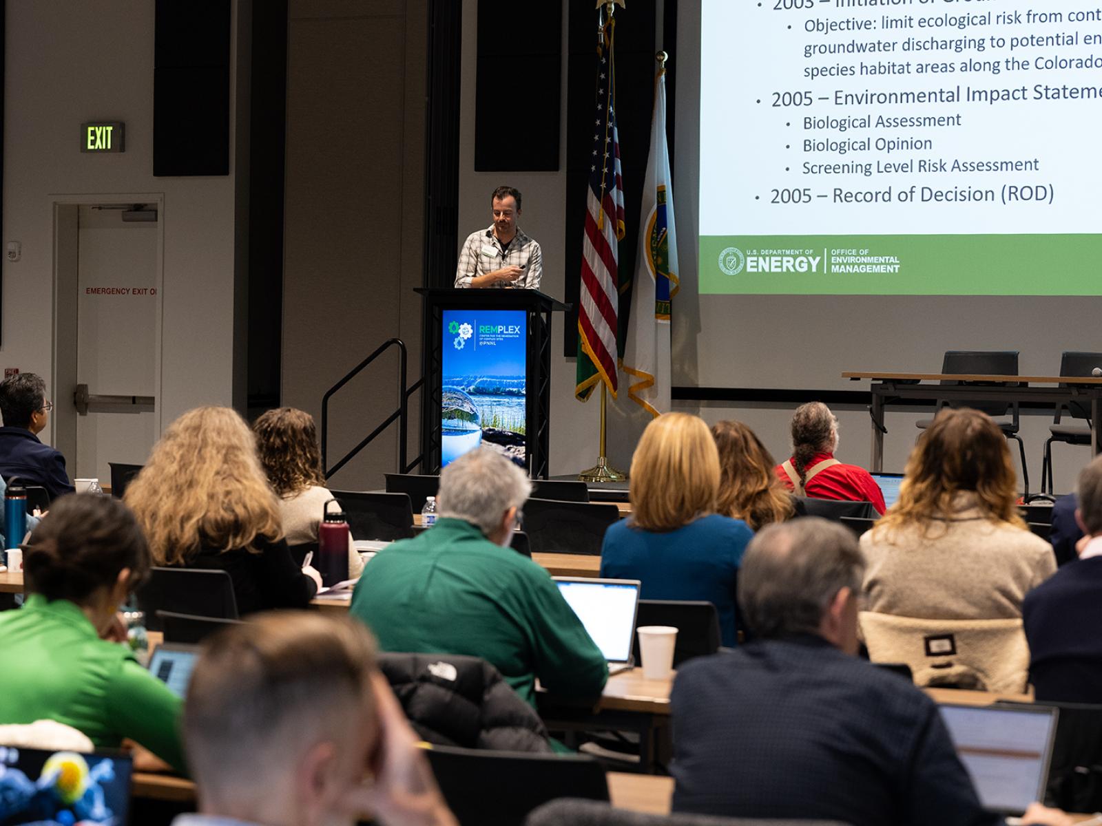 About 100 people sit in a big hall, listening to an environmental remediation expert speaking on stage during the RemPlex Summit